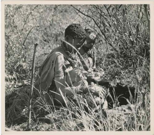 Digging for roots: Woman sitting next to a hole from which she has dug up a root (print is a cropped image)