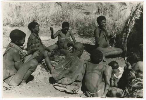 Digging for roots: Group of people sitting in front of Gau's (of Band 2) skerm, including /Gasa, //Khuga (wife of /Naishi), a woman stringing beads, "Little /Qui," and "Old Gau" from Band 1 (print is a cropped image)