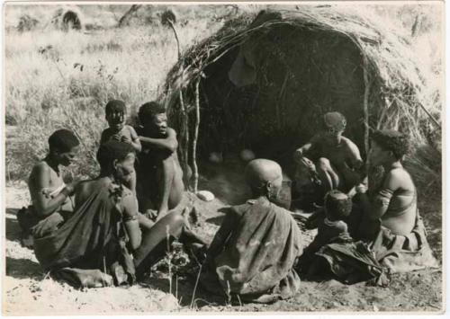 Digging for roots: "/Qui Hunter" cooking and sitting with a group of people in front of a skerm (print is a cropped image)