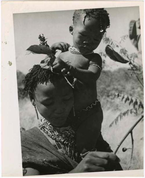 Berry picking: Woman holding a child on her shoulder while she picks berries (print is a cropped image)