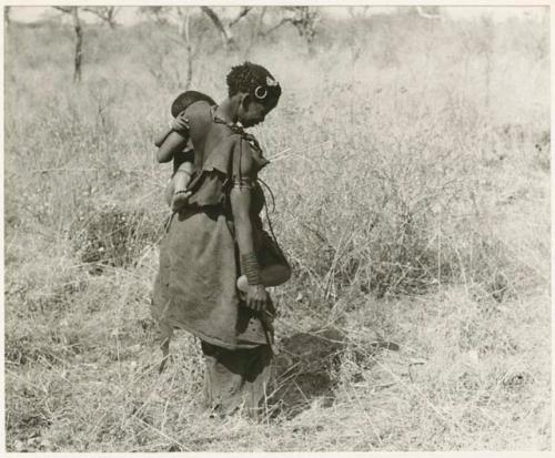 Tsi gathering: Di!ai picking up a stick with her toe in the grass, carrying Debe on her hip (print is a cropped image)