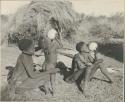 /Gunda (N!ai's husband) steadies a child while the child drinks from an ostrich eggshell, with a woman sitting next to them also drinking from an eggshell