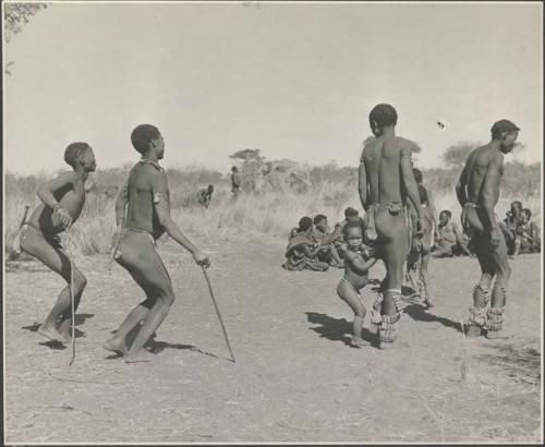 Group of men performing a curing dance, with a child dancing beside her father
