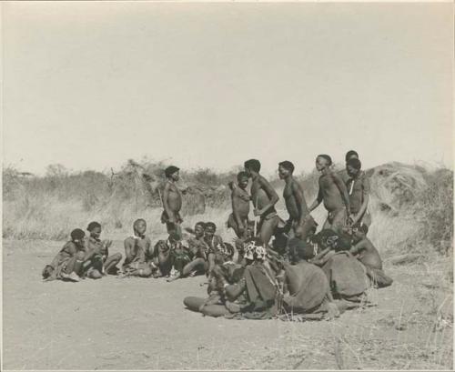 Group of men performing a curing dance, cutting across a circle of women who are clapping and singing