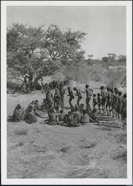 Men and one woman dancing around a circle of women sitting in an early stage of a daytime dance
