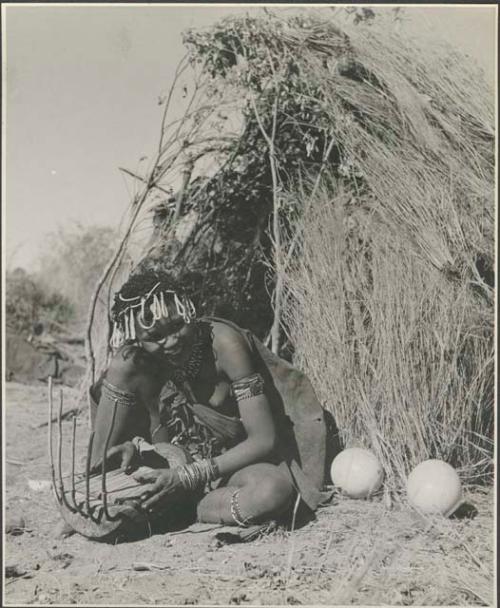 Khuan//a (daughter of !Naishi, headman of Band 5, and //Khuga) sitting in front of a skerm, playing a //guashi
