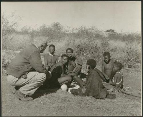 Group of people sitting, including Gau, "Old ≠Toma," /Gam and //Khuaga (wife of !Naishi), with Laurence Marshall crouched next to them
