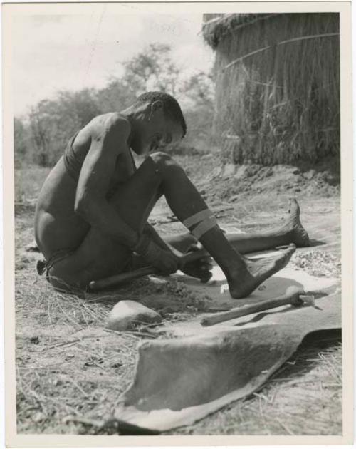 "Scraping hides, working on hides": Man scraping a hide, with his adze lying next to him on the ground (print is a cropped image)