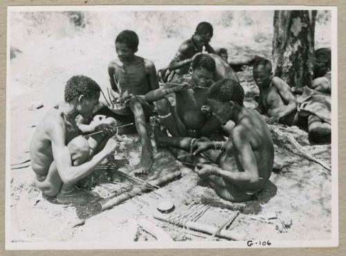 "Scraping hides, working on hides": Group of men working on arrows at the werft of ≠Gao (headman of Band 3), including !Naishi (brother of /Goishay) using sinew to bind the end of an arrow shaft, "/Gao Music" playing the //guashi, and "/Qui Navel" putting poison on an arrow shaft (print is a cropped image)