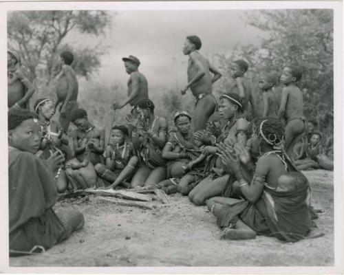 "Ngami in trance, Tsho/ana": Group of women sitting in a circle singing and clapping, with men dancing behind them (print is a cropped image)