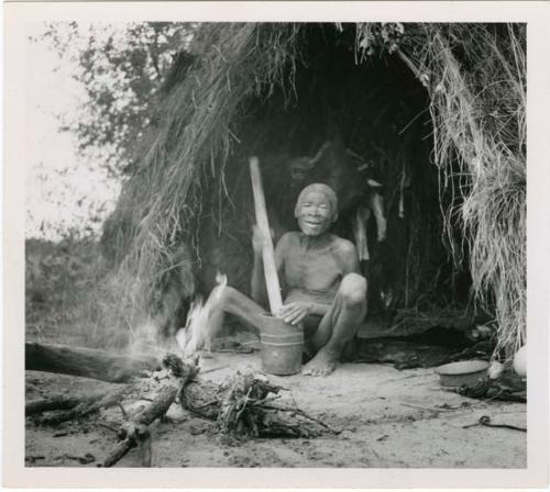 "Mortar and pestle": Man sitting next to a fire in front of his skerm, pounding with a mortar and pestle (print is a cropped image)