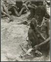 [No folder title]: "Old Xama" preparing a piece of skin, other unidentified people in the background (print is a cropped image)