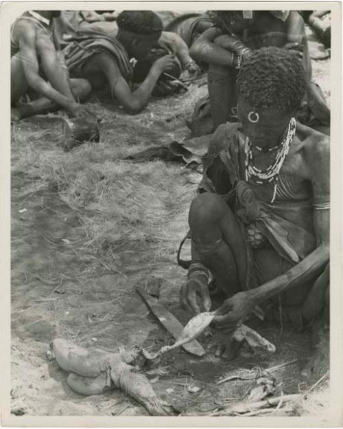 [No folder title]: "Old Xama" preparing a piece of skin, other unidentified people in the background (print is a cropped image)