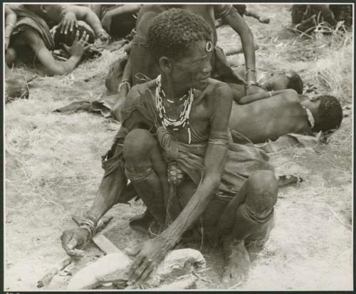 [No folder title]: "Old Xama" preparing a piece of skin, other unidentified people in the background (print is a cropped image)