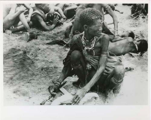 [No folder title]: "Old Xama" preparing a piece of skin, other unidentified people in the background (print is a cropped image)