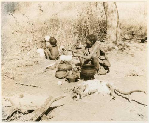 [No folder title]: Child drinking from an ostrich eggshell and a woman (likely /Naoka, U!'s mother) sitting beside a fire with a pot and bones before her (print is a cropped image)