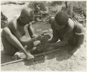 "Firemaking": Dam and ≠Gao creating a fire with firesticks; unidentified people in the background (print is a cropped image)