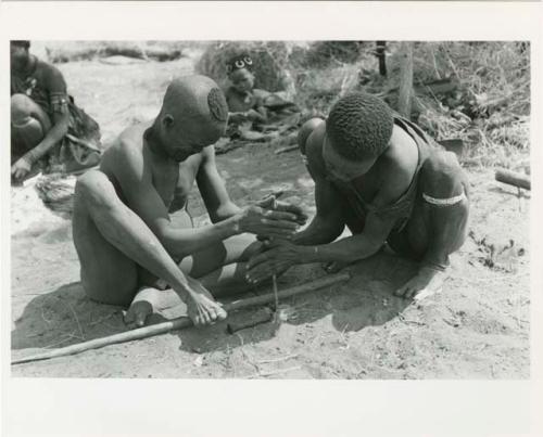 "Firemaking": Dam and ≠Gao creating a fire with firesticks; unidentified people in the background (print is a cropped image)