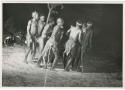 "Night dance at Tsumkwe 1957-8, some published in my [Lorna Marshall's] dance paper": Men dancing in a line, with the man in the center leaning forward (print is a cropped image)