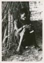 "Night dance at Tsumkwe 1957-8, some published in my [Lorna Marshall's] dance paper": Man wearing dance rattles, sitting by a skerm (print is a cropped image)