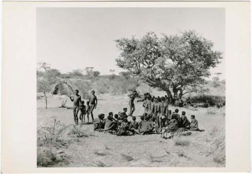 "Daytime dance at Gautscha": Men and two boys dancing through a circle of women sitting, view from the top of an expedition truck (print is a cropped image)