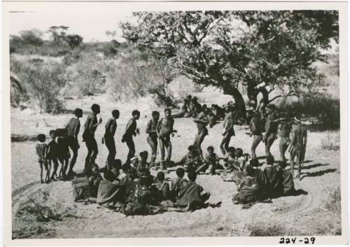 "Daytime dance at Gautscha": Men dancing in a line through a circle of women sitting, with a group of people sitting under a tree in the background, view from the top of an expedition truck (print is a cropped image)