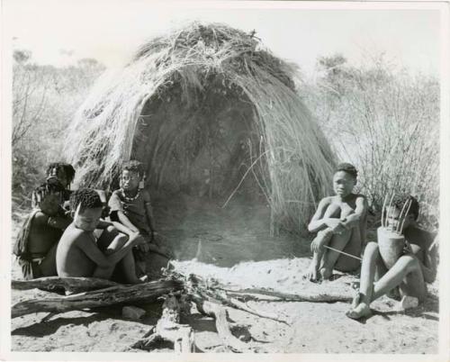 "N!ai and /Gunda": "Little N!ai" and /Gunda (son of Khuan//a and her former husband) sitting by their skerm with their wedding party, including Tsamgao (son of ≠Toma and !U), ≠Gisa (daughter of ≠Gao and Khwo//o-/Gasa), Xama (daughter of "Gao Helmet" and //Kushay), ≠Gao (son of Gau and Be), and a boy playing the //guashi (print is a cropped image)