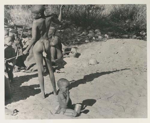 N!whakwe in werft eating from a tin, with empty tsama melon rinds and a group of other boys in the background


