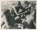 N!whakwe leaning against his father !Gai and holding a turtle, with a group of people around the fire


