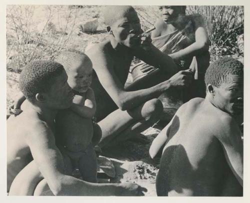 N!whakwe leaning against his father !Gai and holding a turtle, with a group of people around the fire


