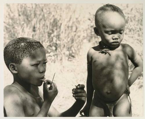 /Gaiamakwe eating a piece of meat, with N!whakwe standing next to him



