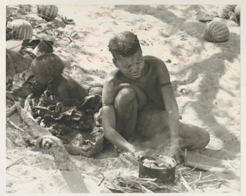 Tsekue cooking a stew of meat and tsama melon pulp at the fire, with N!whakwe beside her


