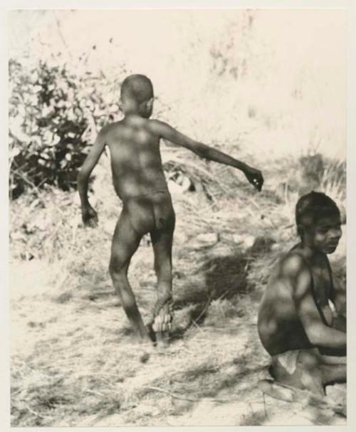 Boy dancing seen from behind, with another boy sitting in the foreground


