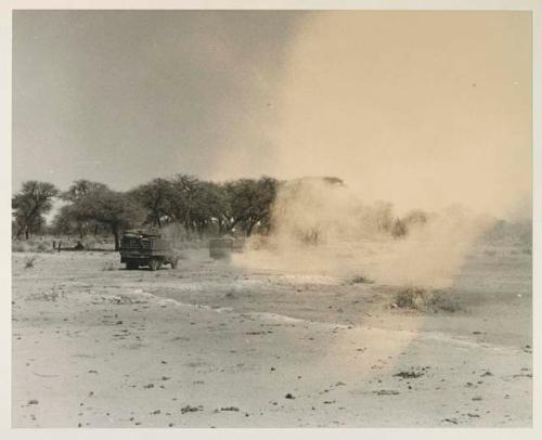 Expedition trucks crossing bare ground, with sand blowing



