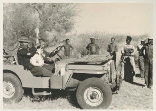 Group of people and Cavasitue standing near Wilhelm Camm sitting in the Jeep holding a guitar, and John Marshall sitting next to him



