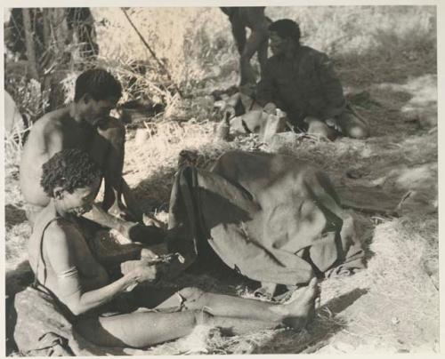 "Gao Medicine" sitting next to a group of children under a kaross, with ≠Toma wearing a coat in the background
