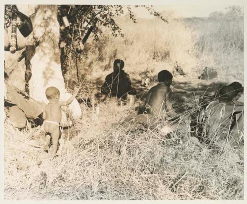 Group of people sitting under a tree, including ≠Toma, and shows belongings hung in the tree
