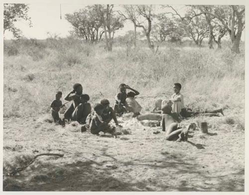 A group of people seen from a distance, sitting in the sun, including !U with !Ungka Norna in her lap, Di!ai, and her son /Gaishay beside her, Debe standing beside !Ungka Norna, an unidentified, visiting girl, !Ungka, and "≠Gao Lame"
