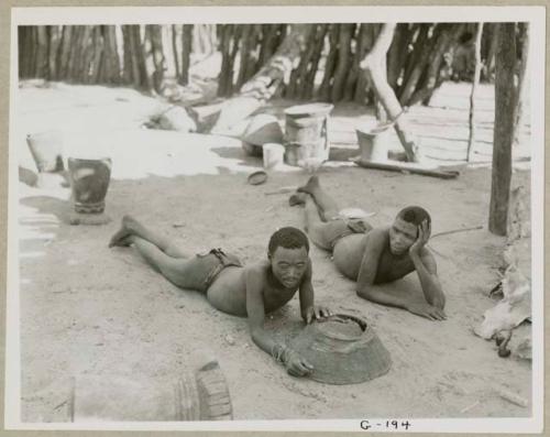 Two men lying on the ground inside Muremi's kraal with grain mortars in the background (print is a cropped image)