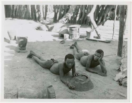 Two men lying on the ground inside Muremi's kraal with grain mortars in the background (print is a cropped image)