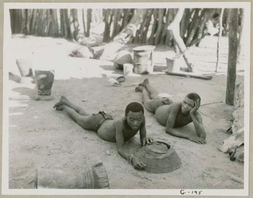 Two men lying on the ground inside Muremi's kraal with grain mortars in the background (print is a cropped image)