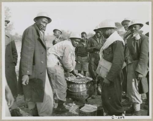 WNLA (Witwatersrand Native Labor Association) cook making mealie porridge, a group of men watching (print is a cropped image)