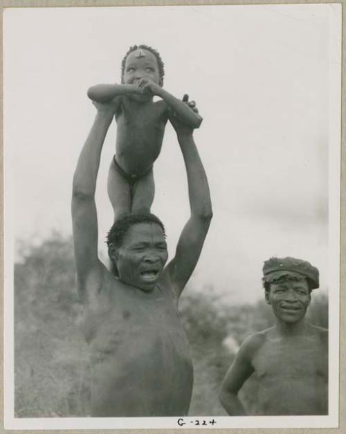 "/Qui Navel" singing medicine songs while holding his son, "Little ≠Gao," on his shoulders and ≠Toma standing to the right wearing a hat (print is a cropped image)