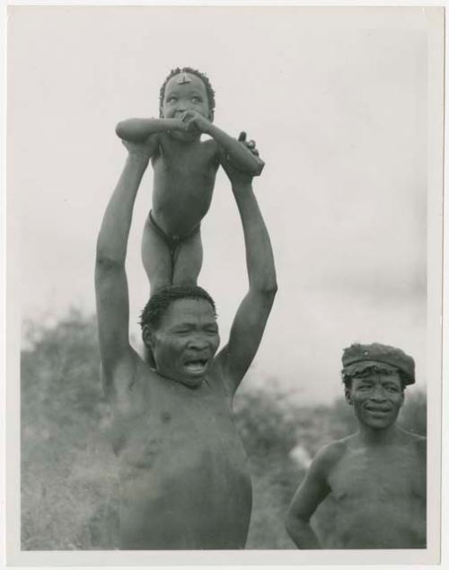 "/Qui Navel" singing medicine songs whole holding his son, "Little ≠Gao," on his shoulders and ≠Toma standing to the right wearing a hat (print is a cropped image)