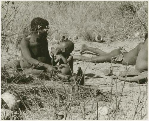 Oukwane sitting with N!whakwe beside him drinking from a tin can (print is a cropped image)