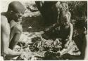 !Gai crouching by his fire cutting meat, N!whakwe and two other people sitting nearby (print is a cropped image)