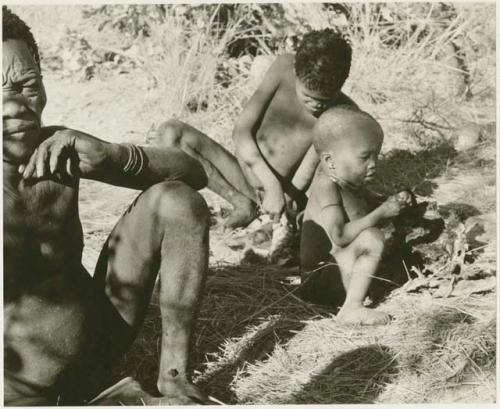 Oukwane sitting, //Kwaiǂkabe cutting meat, N!whakwe sitting nearby (print is a cropped image)