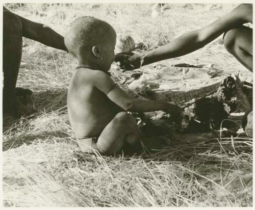 N!whakwe sitting by fire, a person's hand in front of his face (print is a cropped image)