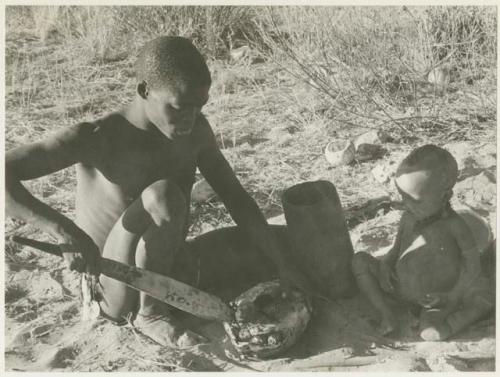 !Gai cutting into a turtle with a fire paddle, N!whakwe sitting beside him (print is a cropped image)