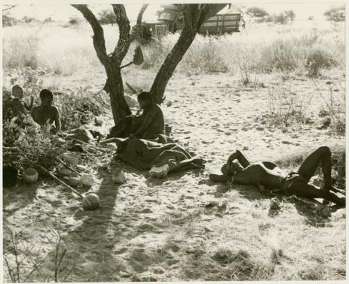 !Gai lying on ground, Tsekue lying under a blanket; /Twikwe, DaSi!Na and N!whakwe sitting nearby (print is a cropped image)